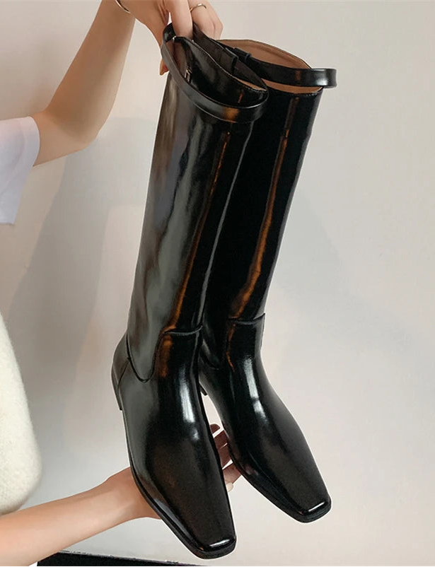 Black glossy knee-high boots being held by a person against a neutral background